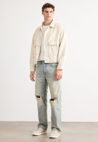 Chaqueta de mezclilla beige con bolsillos frontales, llevada sobre una camisa blanca. Jeans de mezclilla claros y desgastados con desgarros en las rodillas. Zapatillas de colores claros.