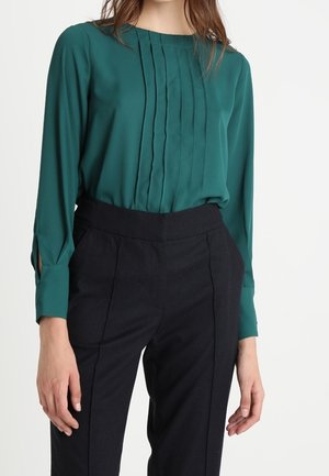 Nainen pukeutuneena vaaleanvihreään pitkähihainen puseroon, jossa pystysuoria laskoksia, ja mustiin korkeavyötäröisiin räätälöityihin housuihin valkoista taustaa vasten.