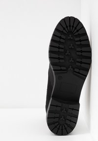 Semelle d'une botte noire avec des motifs de bande de roulement profonds, debout contre un mur d'angle blanc.