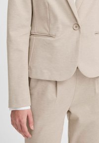 Beige saka s dobře padnoucím střihem, s jedním knoflíkem, předními kapsami a hladkým povrchem. Z rukávu vykukuje bílý košilový manžet.