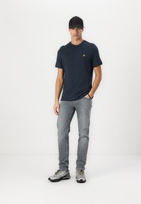 Marineblå t-skjorte med kort erm og gul logo, kombinert med lysegråjeans og grå joggesko. Modellen har på seg en svart caps.