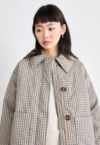 Cappotto a scacchi nelle tonalità beige e blu, con un ampio colletto, grandi tasche e bottoni marroni, realizzato in tessuto testurizzato.