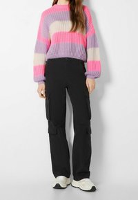 Stickad oversize-tröja i rosafärgad, lila och krämfärgad rand; kombinerad med svarta cargobyxor med sidofickor; vita sneakers.