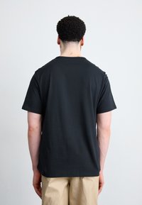 Zwart katoenen T-shirt met korte mouwen en een ronde hals, gladde textuur en een losse pasvorm, gedragen met beige broeken. Alleen achterkant zichtbaar.