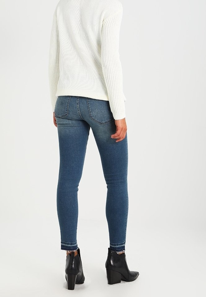 Personne portant un jean skinny bleu, un pull en maille blanc et des bottines noires, debout de dos sur un fond uni.