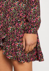 Schwarz geblümtes Kleid mit einem Rüschensaum, das pinke, orange und weiße Blumen zeigt. Das Material scheint leicht und luftig zu sein, mit langen Ärmeln und einem lockeren Schnitt.