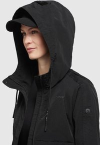 Jachetă neagră cu glugă, realizată dintr-un material texturat. Are fermoare, șnururi și accente cu logo pe piept și umăr. Design casual.