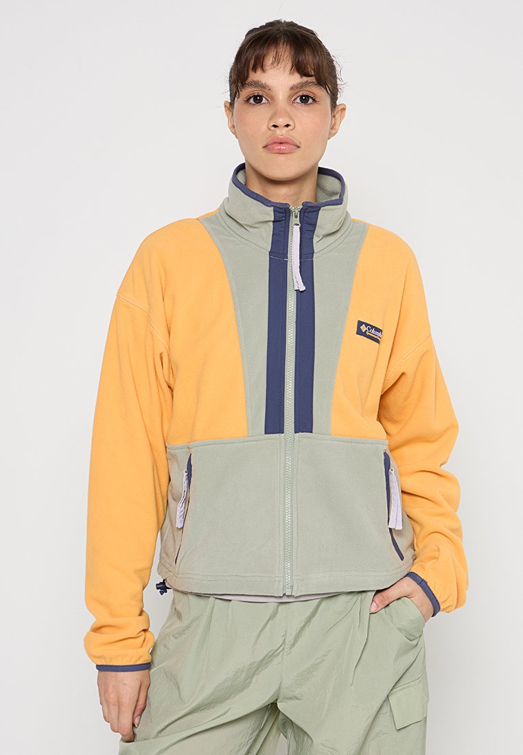 Joven mujer vistiendo una chaqueta de forro polar en bloques de color amarillo y gris con detalles en cremallera azul, de pie con una mano en el bolsillo contra un fondo blanco.