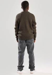 Olivgrüner gerippter Pullover mit Rundhalsausschnitt, kombiniert mit lässigen grauen Jeans. Das Modell steht mit den Händen an den Seiten und blickt von der Kamera weg.
