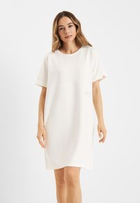 Weißes Kurzarmkleid in entspannter Passform. Hat eine subtile Nahtdetails über der Brust und ein kleines Logo auf der Vorderseite. Glatte Textur.