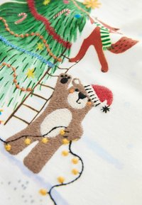 Un tissu brodé présente un ours brun portant un chapeau de Noël, des guirlandes lumineuses suspendues, et un sapin de Noël vert orné de décorations.