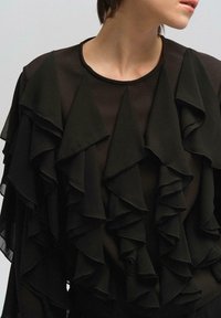 Juoda bluzonė su sluoksniuotais volanais priekyje, apvaliu iškirpimu ir ilgomis rankovėmis. Pagaminta iš lengvo, plazdančio audinio.
