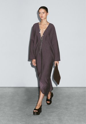 Long purple linen dress with a deep V-neck, oversized sleeves, and side slits. Worn with black platform sandals and a brown clutch.