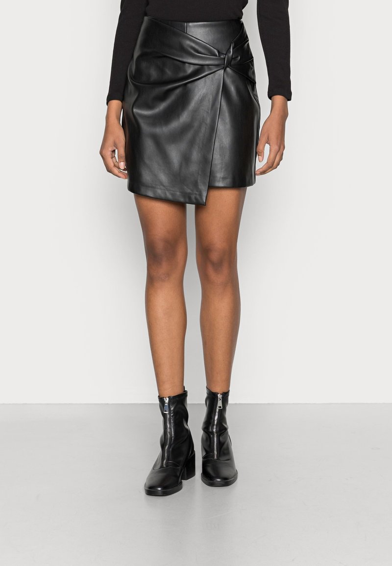 Femme portant une mini-jupe noire en similicuir avec un détail devant qui se chevauche et des bottines noires à fermeture éclair à l'avant.