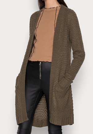 Kvinde iført en tekstureret olivengrøn strikket cardigan over en brun top med sort piping kombineret med sorte bukser med lynlås foran.
