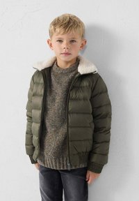 Giacca a vento verde con un morbido colletto in pile bianco, indossata sopra un maglione grigio texturizzato, abbinata a pantaloni grigi scuri.