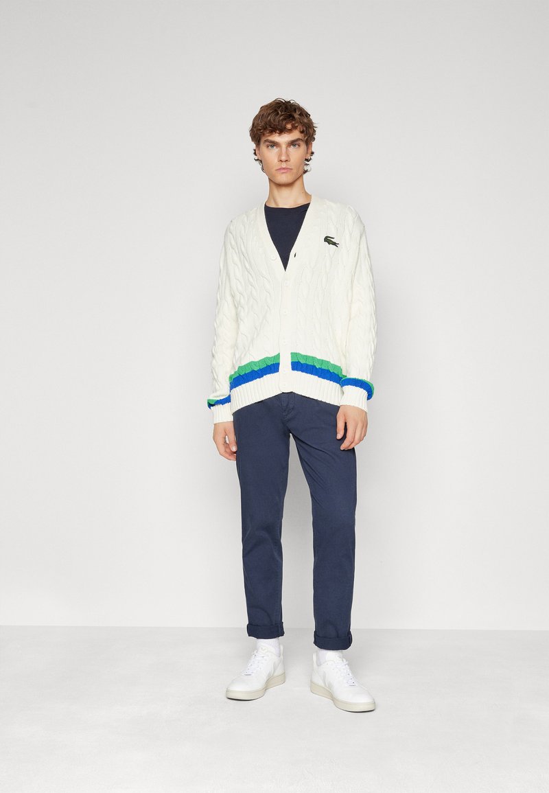 Cardigan en tricot blanc avec des rayures vertes, bleues et marine au bord et aux poignets, porté sur une chemise marine, associé à un pantalon marine et des baskets blanches.