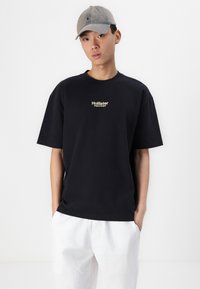 Tricou din bumbac negru, cu mâneci scurte și gât rotund, având un mic logo auriu "Hollister". Asortat cu pantaloni deschiși la culoare.