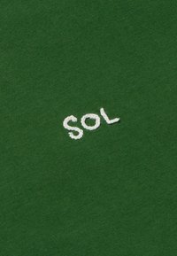 Groene stof met een zachte textuur, waarop in witte borduursel de tekst "SOL" is aangebracht. Het ontwerp is eenvoudig en niet opdringerig.