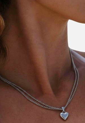 Nahaufnahme des Halses einer Frau, die eine silberne Kette mit herzförmigem Anhänger vor einem neutralen Hintergrund trägt.