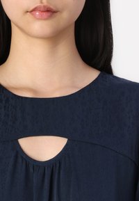 Blusa azul marinho com um tecido texturizado, apresentando um decote redondo e um detalhe em forma de olho de fechadura abaixo do colarinho, com acentos plissados.