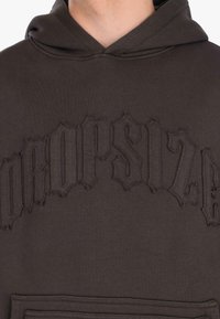 Felpa cappuccio marrone scuro con lettering gotico ricamato in rilievo sul petto e tasca a marsupio frontale.