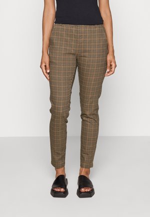 Kobieta ubrana w brązowe, dopasowane spodnie w kratę oraz czarne sandały na płaskiej podeszwie z odkrytymi palcami, stojąca na tle jednolitego, jasnego tła.