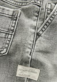 Gros plan sur un tissu en denim gris montrant une poche, des détails de couture, un rivet métallique et une étiquette rectangulaire blanche portant l'inscription « AUTHENTIC GOODS » avec un texte supplémentaire.