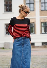 Femme aux cheveux blonds en chignon, portant des lunettes de soleil noires, un pull rayé noir et rouge, et une longue jupe en denim bleu à l'extérieur.