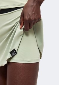 Main à la peau foncée soulevant une jupe vert clair pour révéler un short ajusté assorti avec une étiquette en tissu noire indiquant Endless.