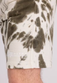 Kratke hlače od pamuka s tie-dye uzorkom u nijansama maslinasto zelene i krem, s bijelim logotipom "ALPHA INDUSTRIES" blizu rubova.