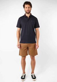 Polo shirt navy avec un motif géométrique, à manches courtes et col, associé à un short beige et des baskets noires avec des semelles blanches.