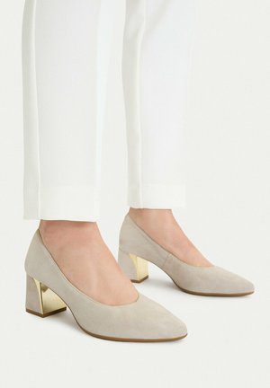 Pies con zapatos de tacón grueso de ante beige con tacones metálicos dorados combinados con pantalones blancos a medida sobre un fondo liso.