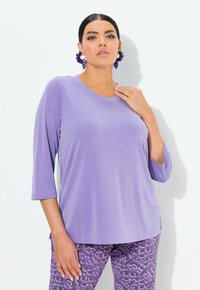 Lila, locker sitzendes Oberteil mit Rundhalsausschnitt und drei Viertelärmeln. Kombiniert mit gemusterten lila Hosen mit geometrischen Designs.