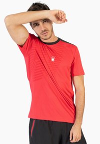 Camiseta atlética roja con un patrón texturizado, cuello negro y un pequeño logotipo de araña blanca en el pecho. Manga corta y tejido ligero.