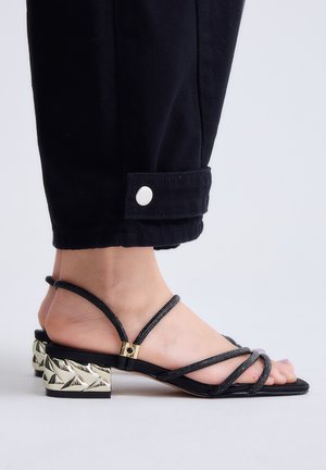 Pie femenino con sandalia negra de tiras y tacón bloque metálico texturizado, combinado con pantalones negros remangados en el tobillo con una lengüeta de botón.