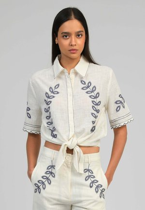 Femme aux longs cheveux foncés portant un ensemble chemise à manches courtes blanche et pantalon avec une broderie de feuilles bleu marine, chemise nouée à l'avant, mains dans les poches.