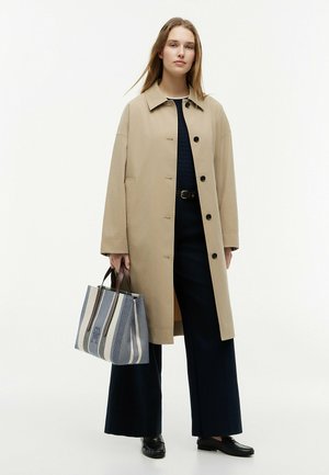 Vrouw draagt een beige lange jas, zwarte wijde broekspijpen en loafers, houdt een blauw-wit gestreepte tas vast.