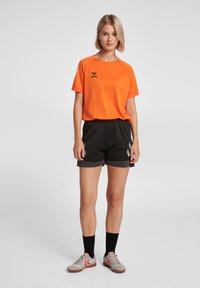 Dames sportoutfit bestaande uit een oranje T-shirt met korte mouwen, zwarte shorts en grijze sneakers met rode accenten. Eenvoudig ontwerp met een merklogo.