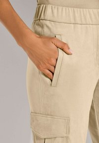 Beige cargo-broek met een zachte textuur, voorzien van een zijzak en een elastische tailleband, benadrukt door een hand die in de zak rust.
