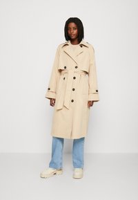 Béžový trench coat s velkými černými knoflíky, širokým límcem a odnímatelným pásem, spárovaný se světle modrými džíny a béžovými chunky platformovými botami.