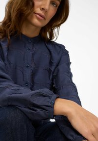 Blusa a maniche lunghe blu navy con motivo ondulato testurizzato, chiusura frontale con bottoni e polsini con dettaglio a bottoni, realizzata in tessuto leggero.