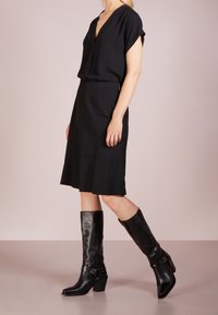 Kvinna i en svart kortärmad blus, svart knälång kjol och svarta läderstövlar med blockklack som når till vaden, står på sidan.