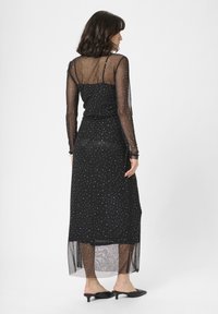 Rochie neagră cu mâneci lungi din material transparent și un corp ajustat. Prezintă puncte albe dispersate și o deschidere laterală, asortată cu pantofi negri slip-on.