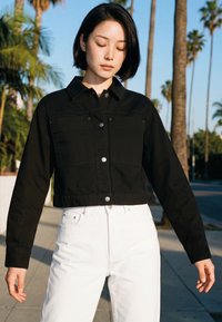 Fiatal nő fekete bob frizurával, fekete gombos kabátot és fehér nadrágot viselve, szabadtéren áll, pálmafák háttérrel.