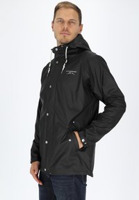 Sort vandtæt jakke med hætte, trykknapper, snøre og logodetalje på brystet. Model viser sidelommer.