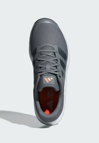 Zapatilla deportiva gris con parte superior de malla texturizada, suela de goma y plantilla naranja. Presenta tres rayas negras y un acento de logo. Diseño con cordones.