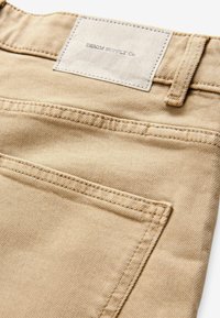 Bežové denimové nohavice s textúrovaným povrchom, s obdĺžnikovou textilnou etiketou, na ktorej je napísané "DENIM SUPPLY Co." a viditeľnými švíkmi.