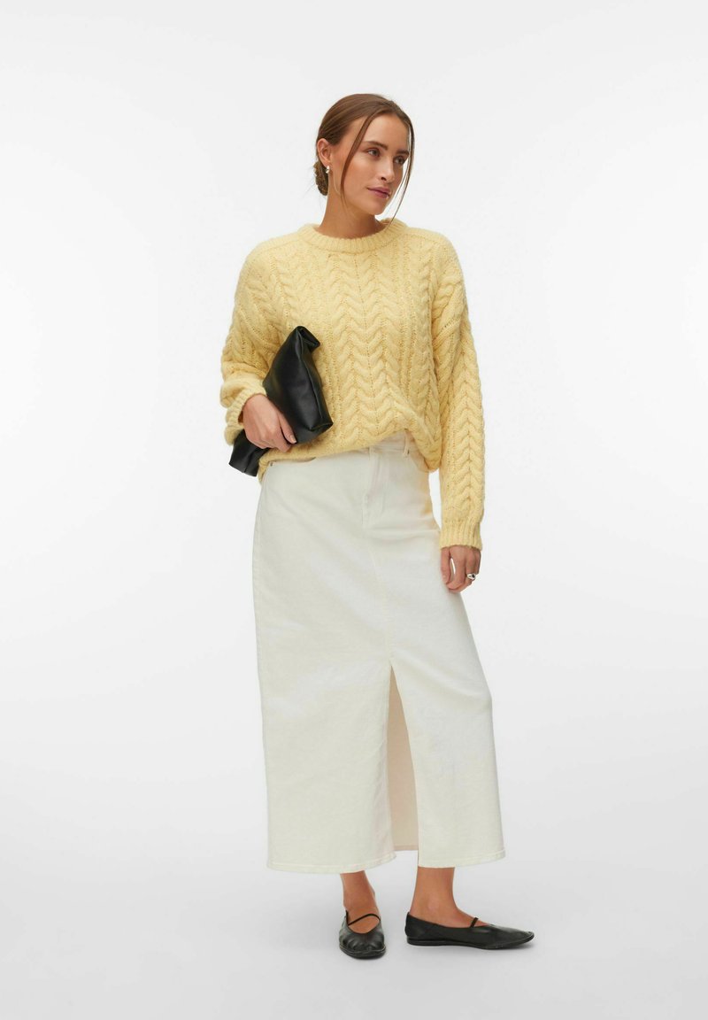 Femme portant un pull jaune en maille torsadée, une jupe midi blanche fendue à l'avant, des ballerines noires, et tenant une pochette noire.