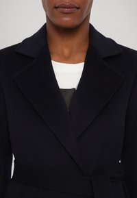 Vlněný kabát v námořnické modři s klopovým límcem, páskem v pase a jemnou texturou. Nošený přes bílou košili s grafickým potiskem pod ní.